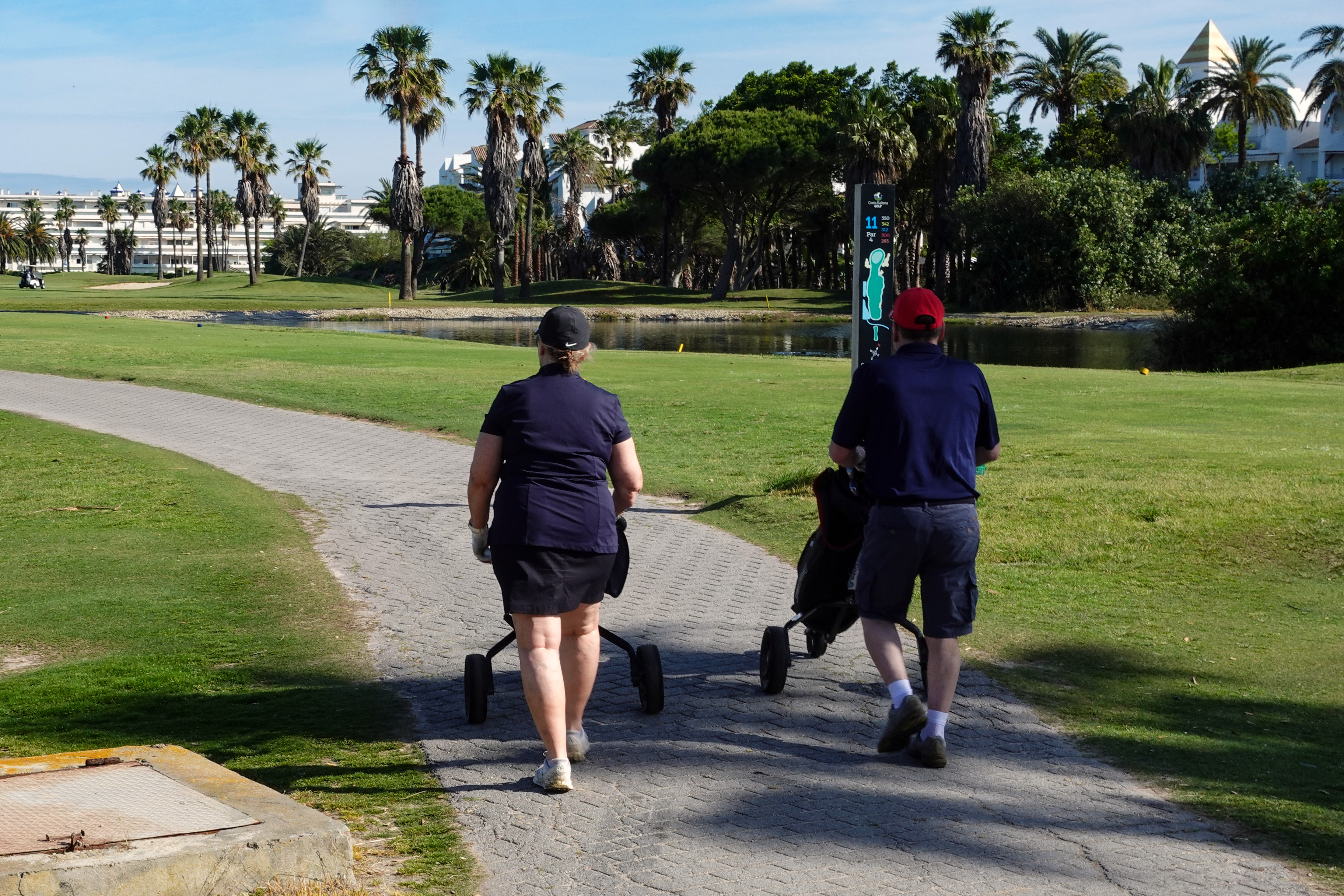 Two travelers walking on a sunny golf course path while pushing wheeled golf carts
